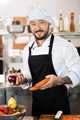 Positive asian chef holding organic vegetables near cutting board in kitchen