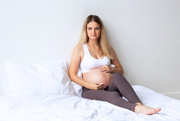 Cute blonde pregnant girl sitting on the bed at home. On a light background. Portrait. 8 month of pregnancy. The concept of waiting for the birth of a baby. Copyspace.