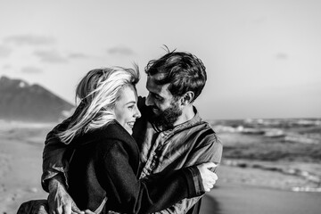 Romantic happy couple in love hugging each other on shoreline at the wild beach - Boyfriend and girlfriend smiling in front of the sea during a big hug - Black and white