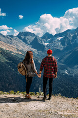 Fototapeta na wymiar A stylish couple in the mountains admiring the beautiful views. A man and a woman with backpacks on the background of mountains. A traveling couple in the mountains. Hiking in the mountains.