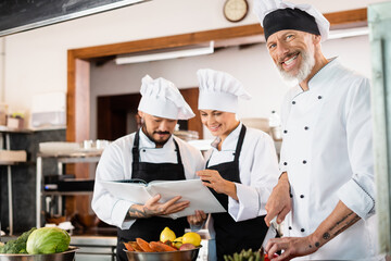 Positive chef holding knife near multiethnic colleagues in aprons reading cookbook and vegetables in kitchen