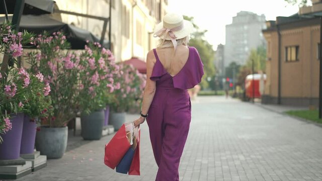 Live camera follows happy relaxed slim woman strolling with shopping bags on beautiful city street. Back view confident Caucasian shopper walking with purchases outdoors. Consumerism and femininity