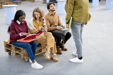 Fototapeta premium Multiracial students reading books while sitting on wooden bench outdoors at day. Concept of education and learning. Idea of student lifestyle. Young modern guys and girls at university campus