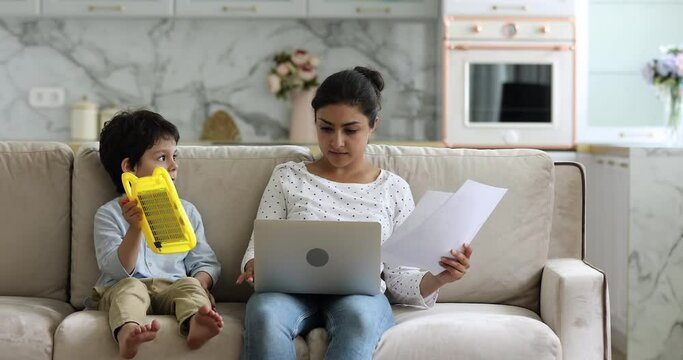 Busy Indian mom work from home with papers laptop praise with smile little son showing new picture drawn on dry erase board. Young mum freelancer engaged in work sit on sofa pay attention to small kid