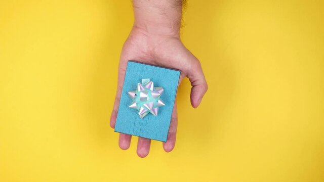 A man's hand holds a blue gift box with a shiny bow on a yellow background