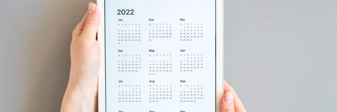 tablet computer with an open app of calendar for 2022 year in a womans hands on a gray background. concept business or to do list goals with technology using. top view, flat lay. banner