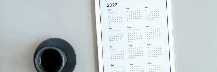tablet computer with an open app of calendar for 2022 year and cup of tea or coffee on a gray background. concept business or to do list goals with technology using. top view, flat lay. banner