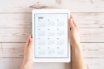 Obraz premium tablet computer with an open app of calendar for 2022 year in a womans hands on a wooden boards background. concept business or to do list goals with technology using. top view, flat lay