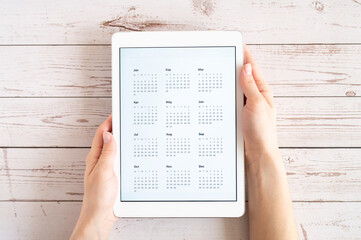 Obraz premium tablet computer with an open app of calendar for unspecified unknown date year without date in a womans hands on a wooden boards background. concept business or to do list goals. top view, flat lay