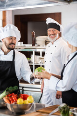 Positive chef looking at interracial colleagues with vegetables in kitchen