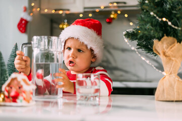 Happy funny toddler boy in a red Santa hat drinking filtered water from a glass in the kitchen. Holidays, health concept.