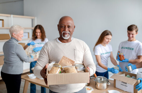 Charity center. Happy black senior man holding box with donations food, looking and smiling at camera