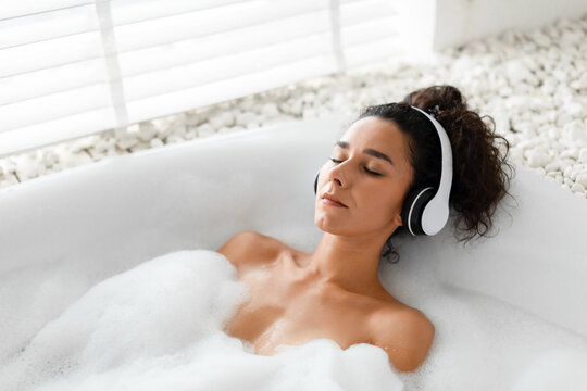 Relaxed young woman listening to calming music in wireless headphones, lying in bubble bath at home, copy space