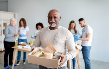 African american senior man holding box with donations food, looking and smiling at camera in...