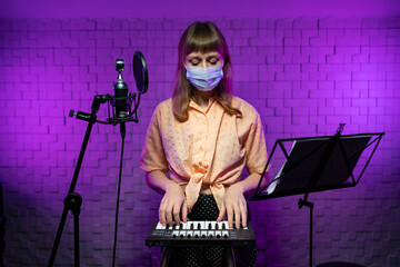 Fototapeta na wymiar Live concert during the pandemic. Caucasian singer female in a protective disposable mask plays the synthesizer, sings into the microphone with a pop filter on stage with a music stand in purple light