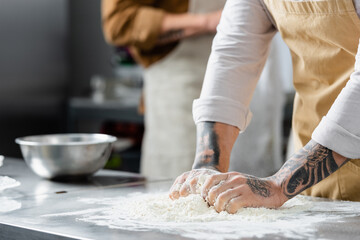 Obraz na płótnie Canvas Cropped view of tattooed chef making dough near blurred colleague in kitchen