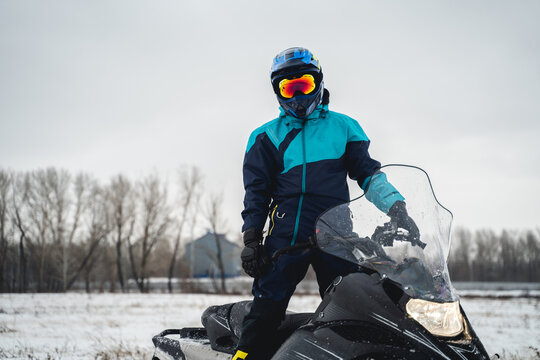 Snowmobile Driver Stands On His Black Snowmobile In A Snowy Area, Cross Helmet