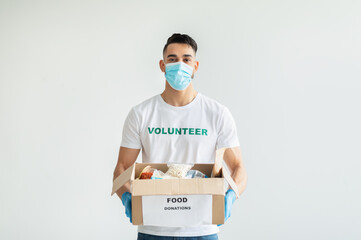 Volunteer work. Young arab man in volunteer t-shirt and medical mask, holding food donations box over light background