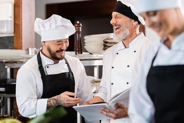 Happy interracial chefs talking while holding cookbook near blurred colleague in kitchen