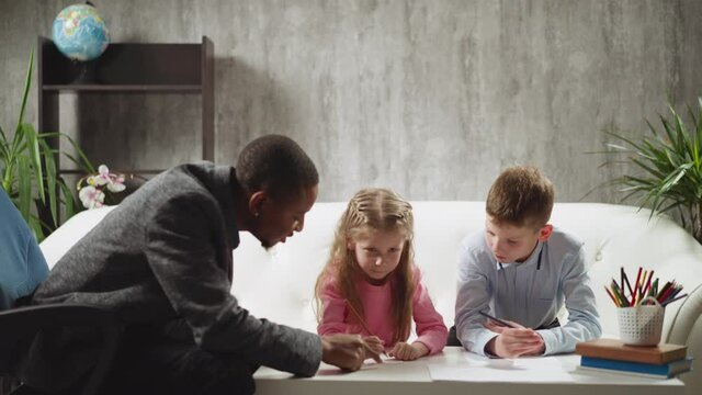 Careful African-American teacher writes letters by finger on table and children try to understand studying at home lesson with skilled tutor slow motion
