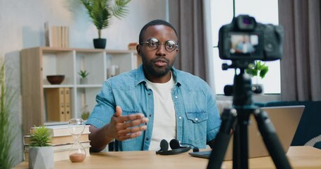 Attractive confident friendly african american blogger in glasses recording video on camera for his internet audience from cozy home office,front view