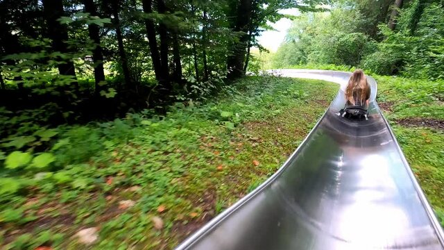 A blonde girl with long hair is riding on a summer toboggan run through a tunnel. Behind the tunnel, beautiful mountains and a blue lake are unfolding.