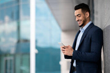 Fototapeta premium Handsome middle eastern businessman standing by business center, using cellphone