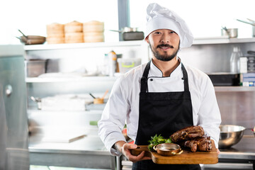 Smiling asian chef holding cutting board with roasted meat and greens in kitchen - Powered by Adobe