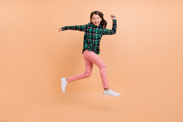 Fototapeta na wymiar Photo of sweet funky school girl dressed checkered clothes smiling running fast jumping high isolated beige color background