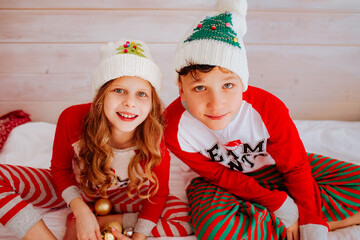Girl and teen boy in Christmas pyjamas and winter hats sitting on the bed