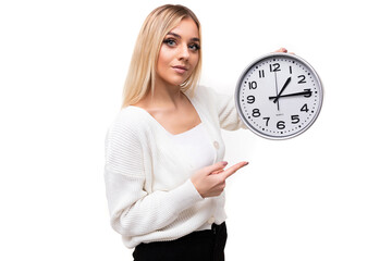 Photo of a beautiful pretty amazing positive young blonde woman in white knitted sweater and black pants points her finger at the big clock isolated on a white background with copy space for text