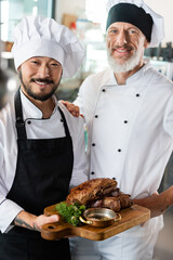 Positive multiethnic chefs holding cooked meat on cutting board in kitchen