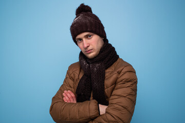 Sad young man in brown scarf and winter hat, light brown jacket on blue background