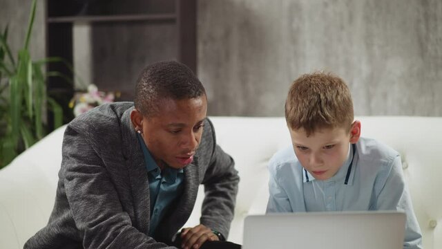 Black native speaker English teacher translates movie to little boy pupil during private tuition via modern laptop at home closeup slow motion