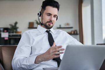Businessman wearing headset sitting at the armchair and gesturing while having video call
