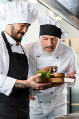 Mature chef smelling roasted meat near asian colleague in kitchen