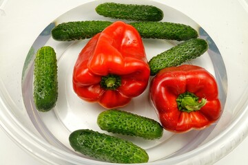 Red bell peppers and fresh cucumbers are in a plastic bowl with water. Washing fruit before eating.