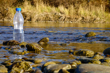 Obraz na płótnie Canvas A plastic bottle with drinking water stands on a stone in the river against the background of an autumn grove