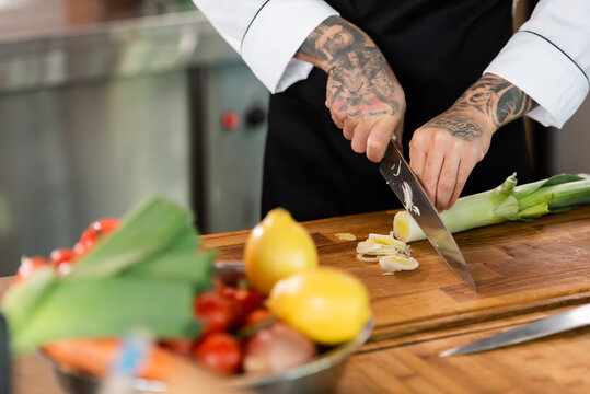 Cropped view of tattooed chef cutting leek near blurred vegetables in kitchen