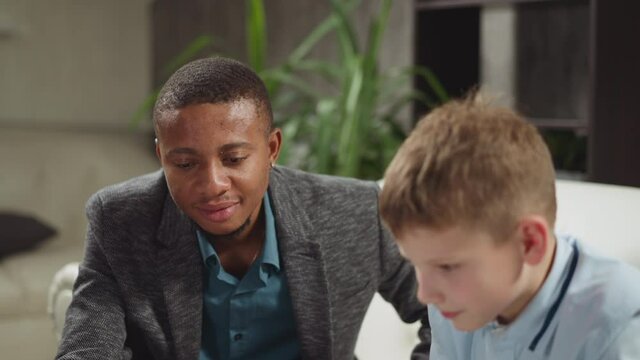 Smiling black guy tutor supports attentive junior schoolboy at private English lesson with native speaker in living room closeup slow motion