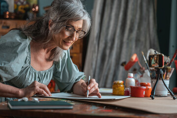 Woman looking at her painting at home while drawing with pencil in front of the smartphone