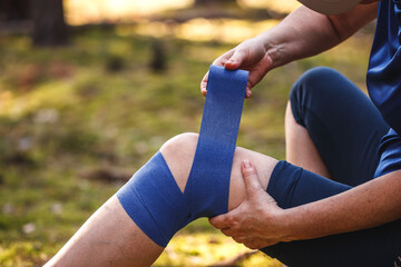 First aid after hiking accident. Injured hiker putting elastic bandage to her knee. Tendon problems or sprain knee during trekking in nature - obrazy, fototapety, plakaty