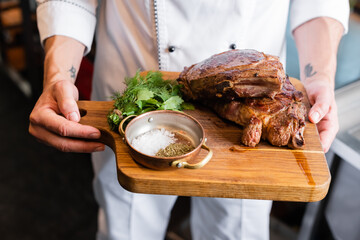 Cropped view of chef in uniform holding cutting board with spices and roasted meat
