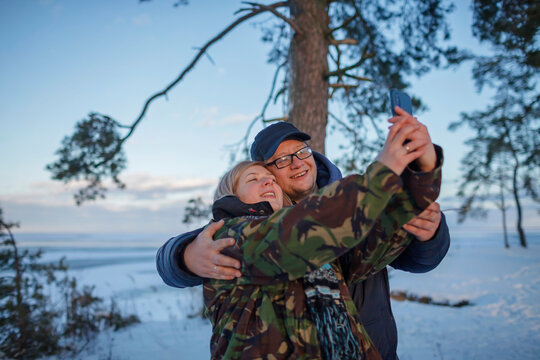 Middle aged man and woman in military jacket with camouflage pattern look at smartphone camera to make selfie, reunion of couple on the shore of beautiful frozen lake in winter