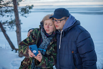 Middle aged man and woman in military jacket with camouflage pattern look at smartphone camera to make selfie, reunion of couple on the shore of beautiful frozen lake in winter