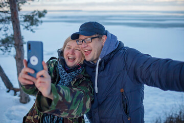 Reunion of couple on the shore of beautiful frozen lake in winter. Middle aged man and woman in military jacket with camouflage pattern look at smartphone camera to make selfie