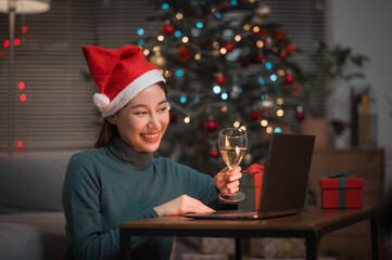 Asian woman wearing Christmas hat drinking champagne video call celebrate Happy New Year party online with her friends or family via laptop at home