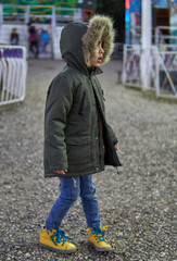 latino little boy dressed in jeans and parka in fur hood lost in an amusement park, walking alone. wintertime, vertical - obrazy, fototapety, plakaty