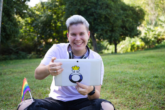 non-binary person sitting in the park with a tablet with the non-binary symbol and the gay pride flag on his knee. Diversity and gay pride. Non-binary visibility concept.