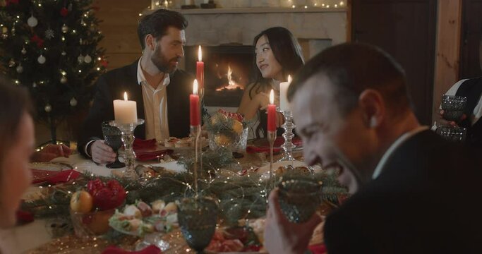 Cheerful Group of Friends in Festive clothes, Having Christmas Dinner Table, sitting in Restaurant with Fireplace in Cozy Atmosphere 